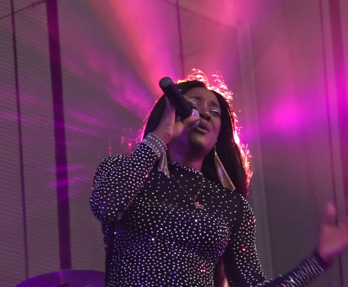 Singer performing onstage under purple lights, holding a microphone and wearing a sparkly black outfit
