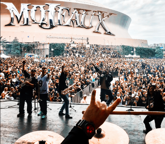 Concert crowd and performers on stage in front of a large “Mojaxx” sign, viewed from behind a drum set