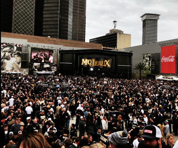 Crowd at an outdoor concert with a large stage and city buildings in the background