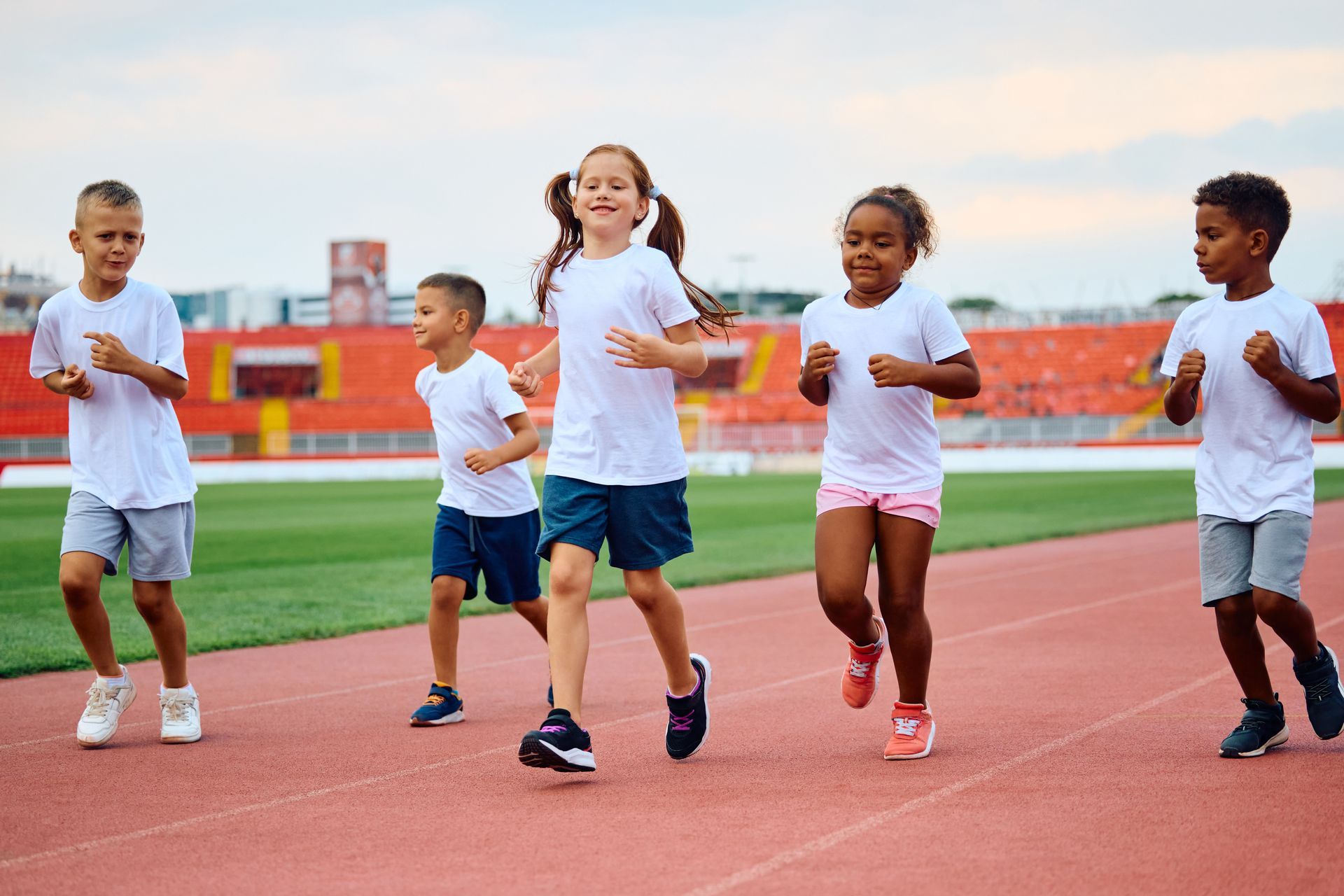 Children running on a track, wearing white shirts and shorts; smiling. Stadium in the background.