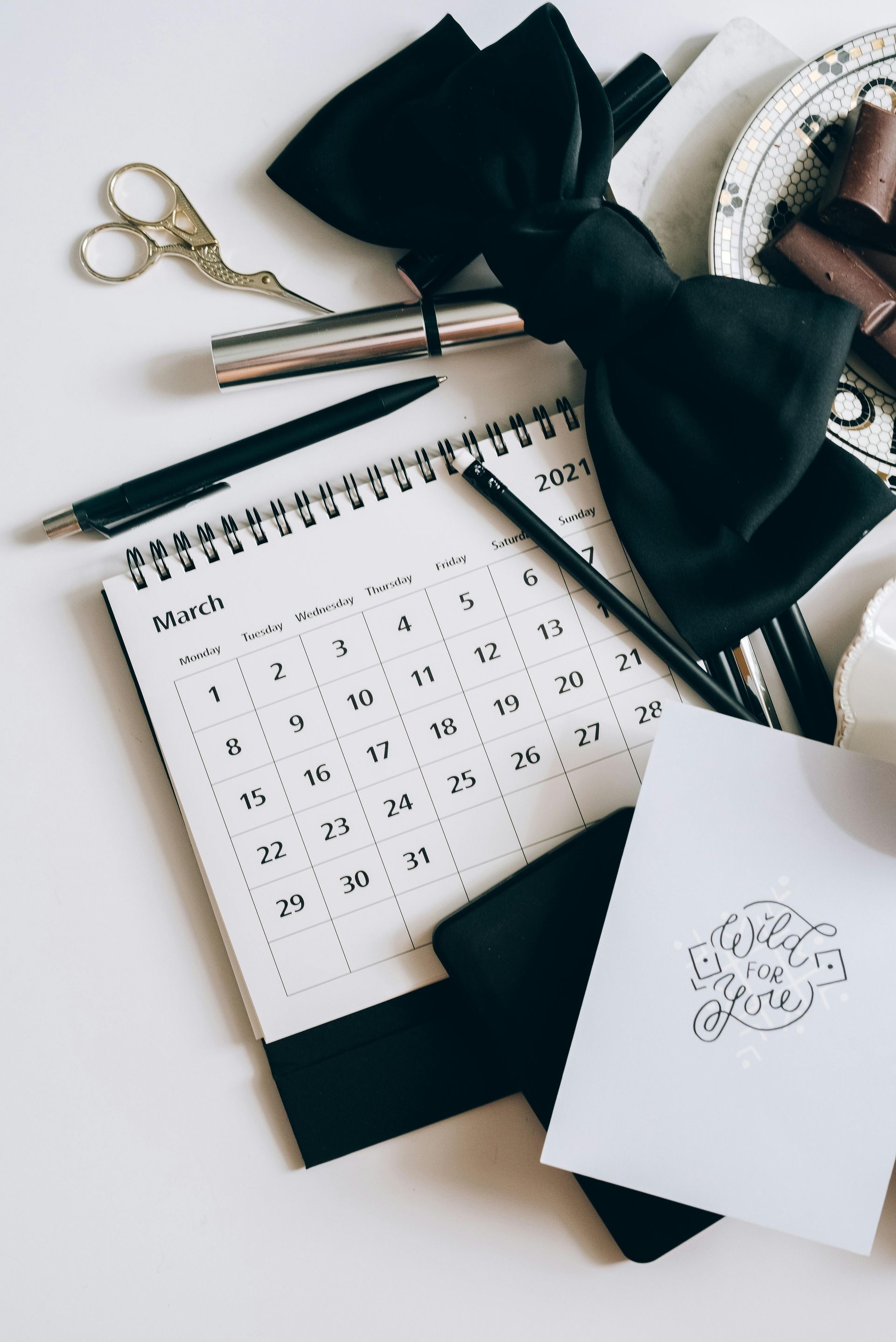 March calendar, black bowtie, pens, scissors, lipsticks, and cards on a white surface.