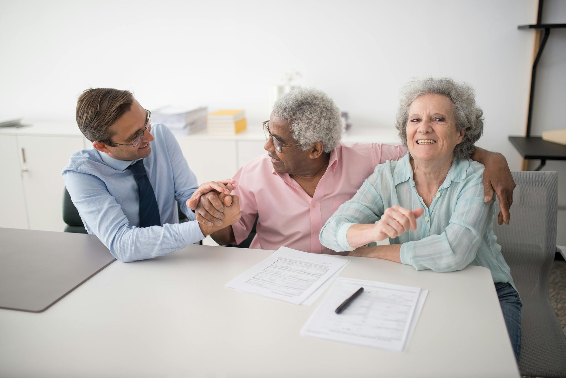A man and a couple smiling around a table; the man holds their hands, the woman has an arm around the man.