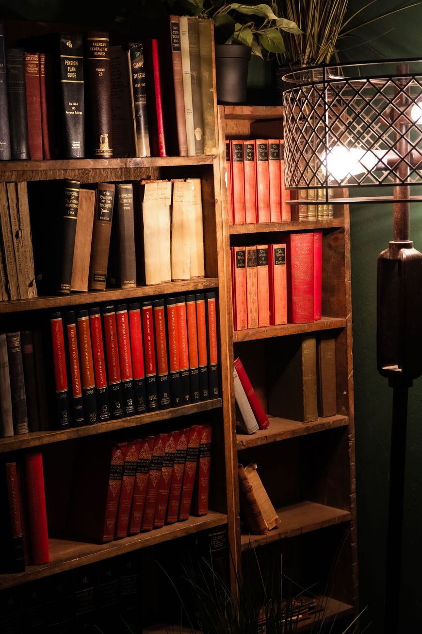 Bookshelves filled with red and black books beside a lit lamp in a cozy, dim study.