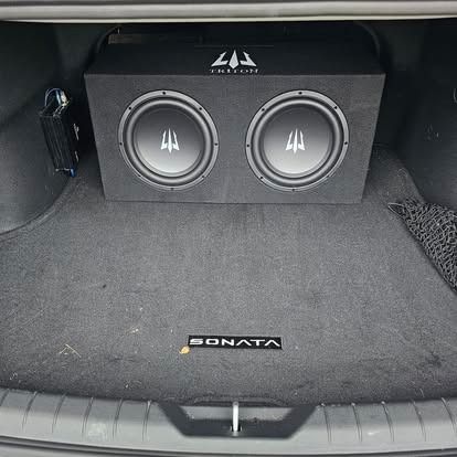 Black car trunk with two subwoofers and a car audio setup behind a DMTA floor mat