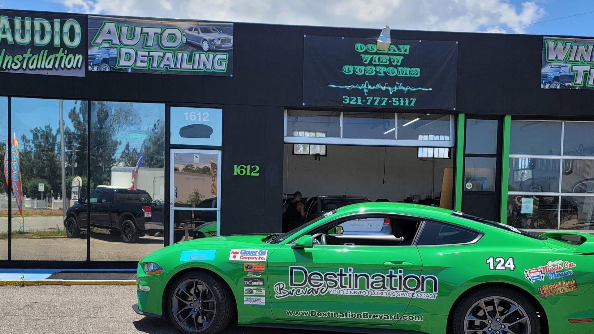 Bright green race car parked outside an audio and auto detailing shop storefront.