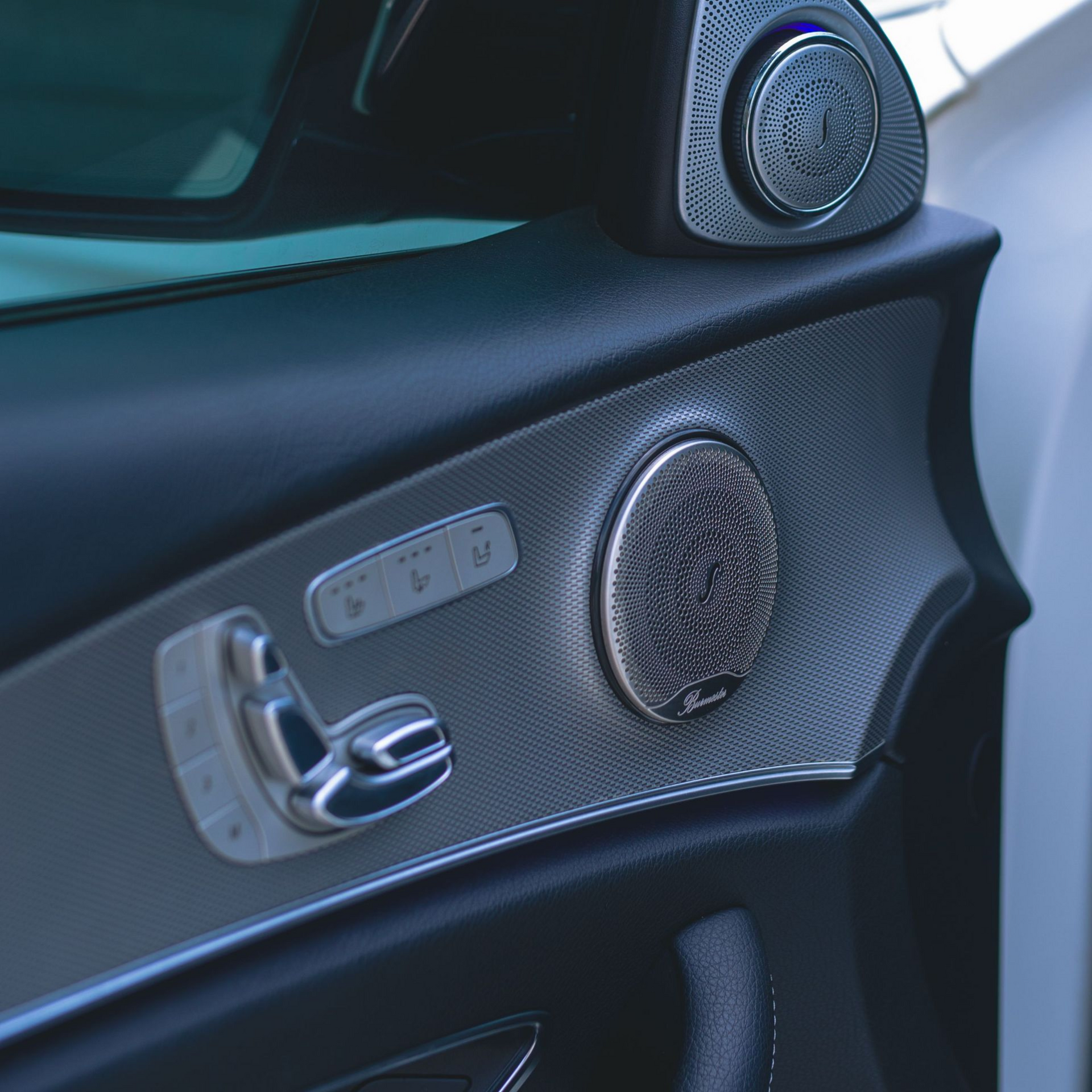 Close-up of a car door interior with silver handle, control buttons, and round speaker grilles.