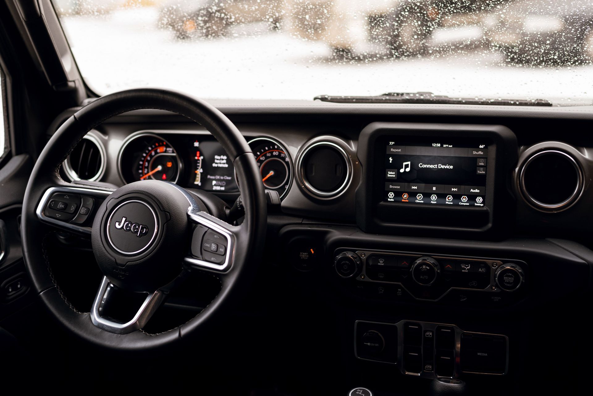 Car interior dashboard and steering wheel with digital display and round vents, seen through a rainy windshield