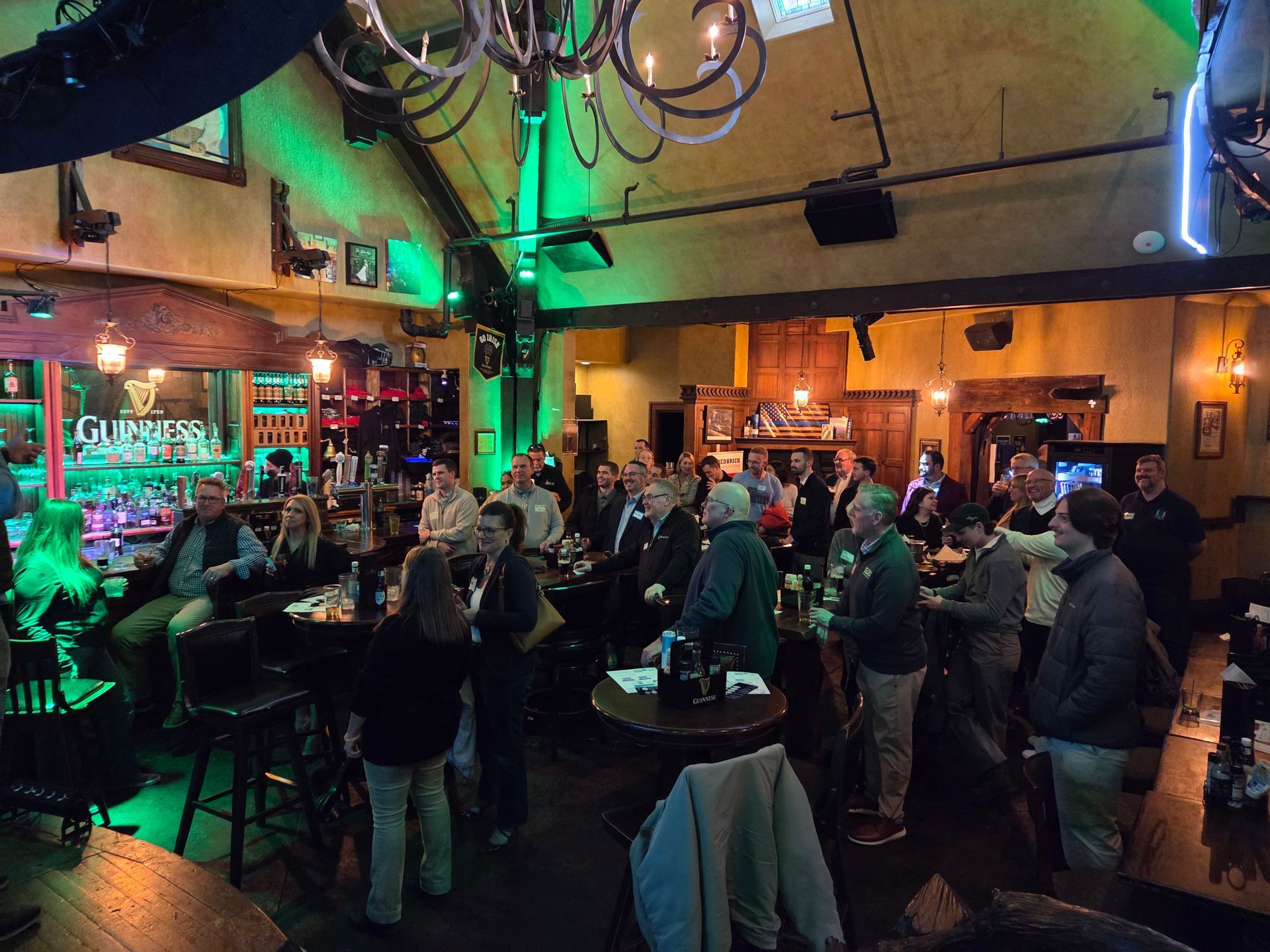 People socializing in a bar; some at the counter, others standing. Lit bar area with drinks and bottles.
