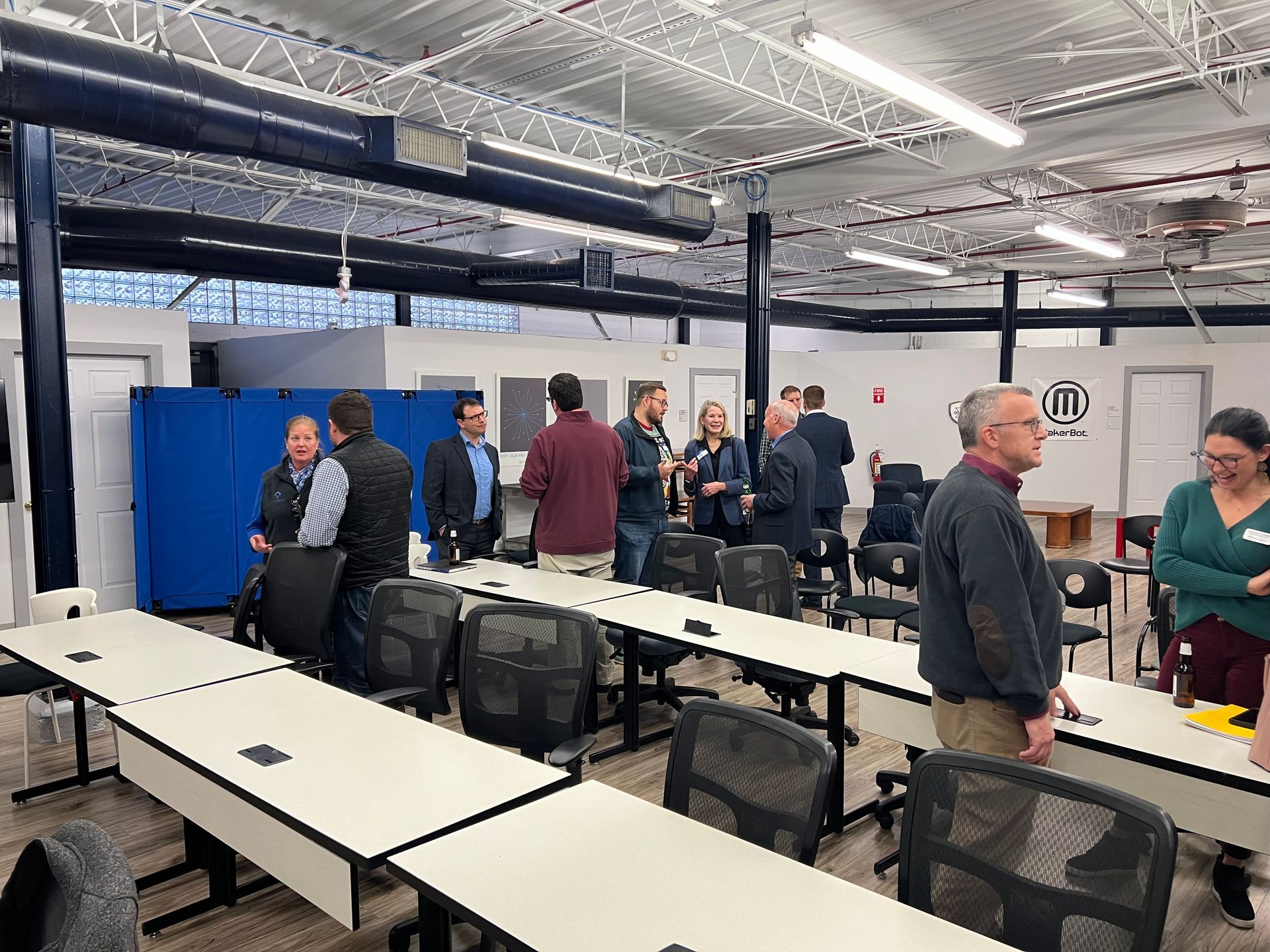 People gather in a large room with long tables and chairs. Some are talking in small groups. Exposed ceiling, blue panels.