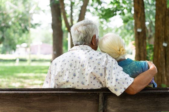Two elderly people sit close together on a bench in a park, viewed from behind.