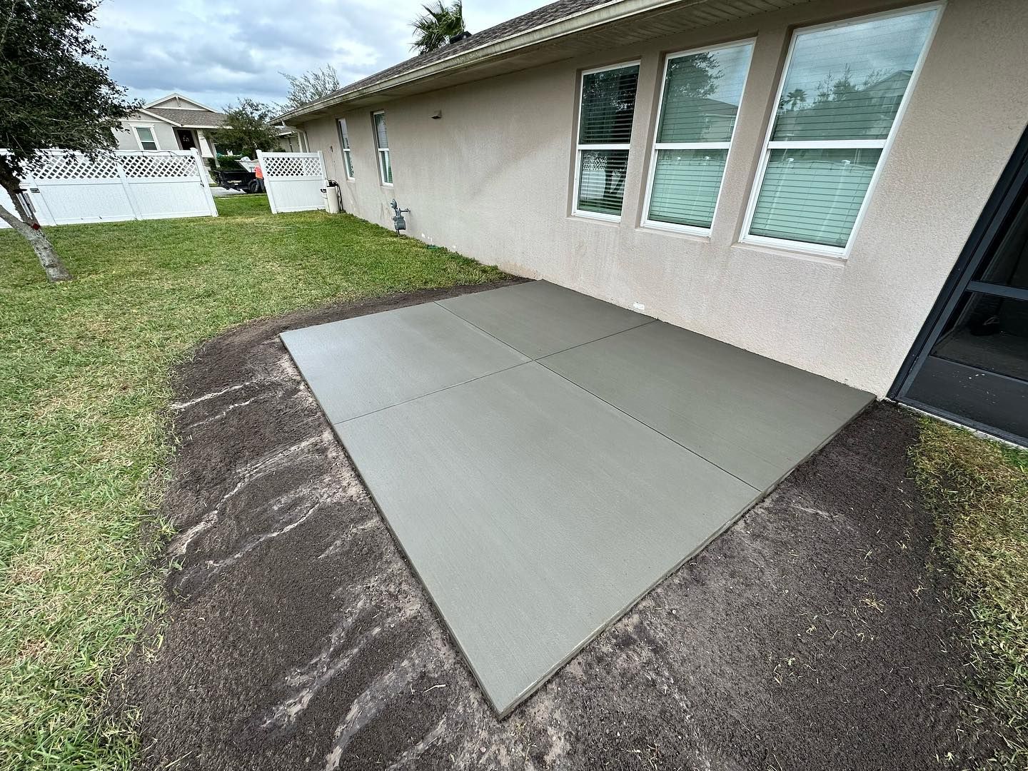 A small grey patio next to a house with windows, surrounded by dirt and grass.