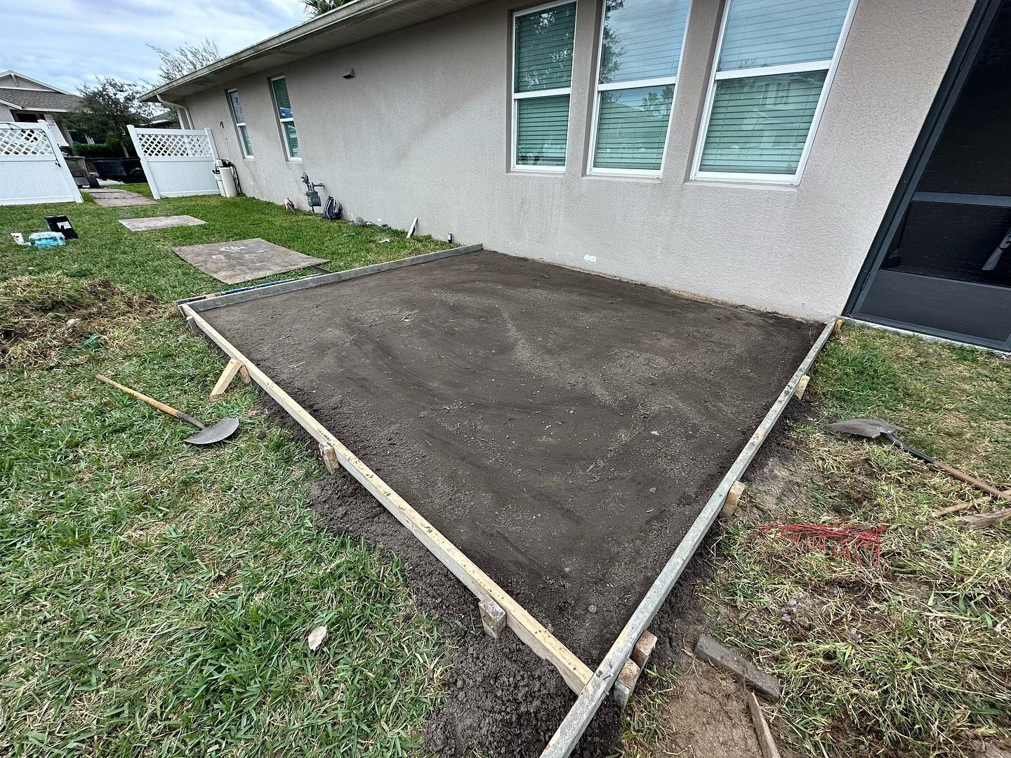 A rectangular wooden frame holds packed dirt for a new concrete patio next to a house.