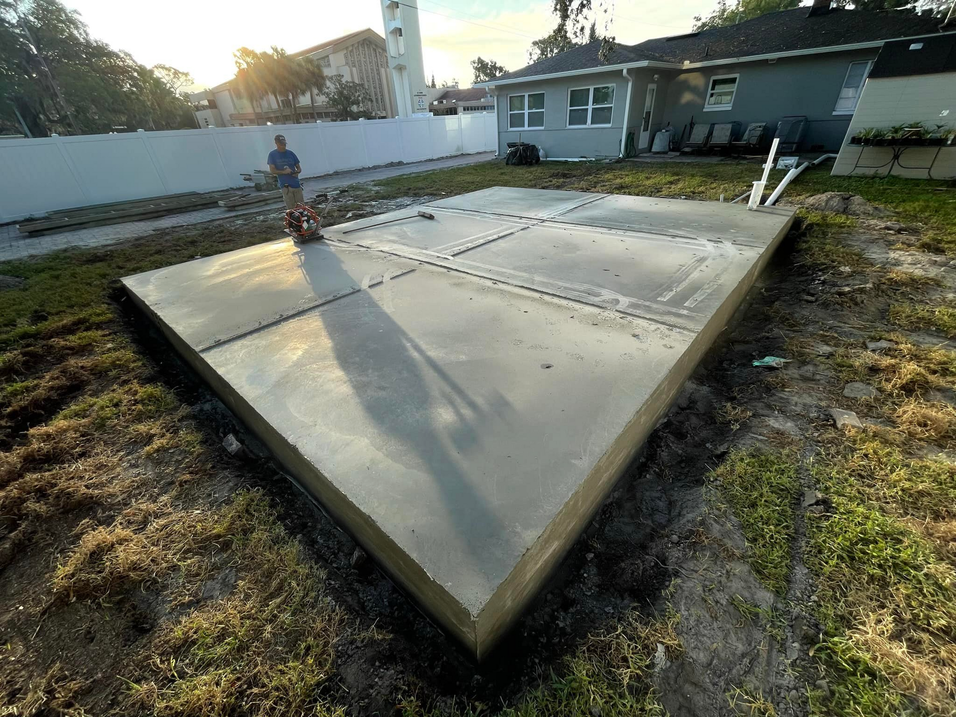 Newly poured concrete slab in a backyard. A person works in the background near a house.