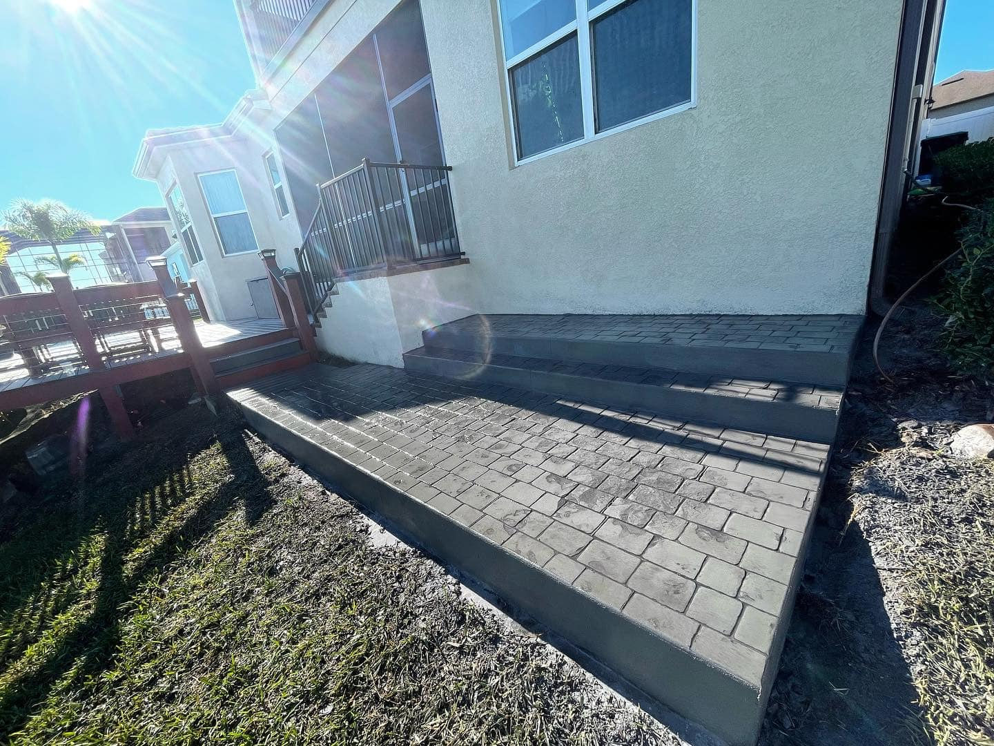Outdoor patio with gray pavers and raised border, next to a beige house, sunny day.