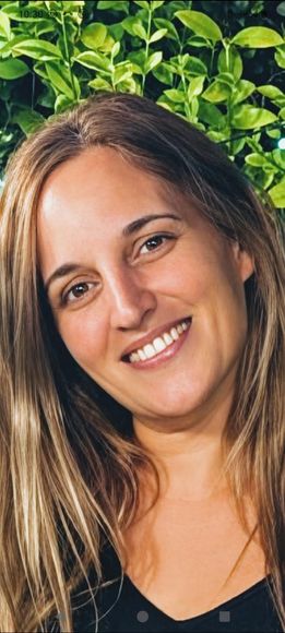 Mujer sonriente de pelo largo con blusa negra, de pie frente a un follaje verde.