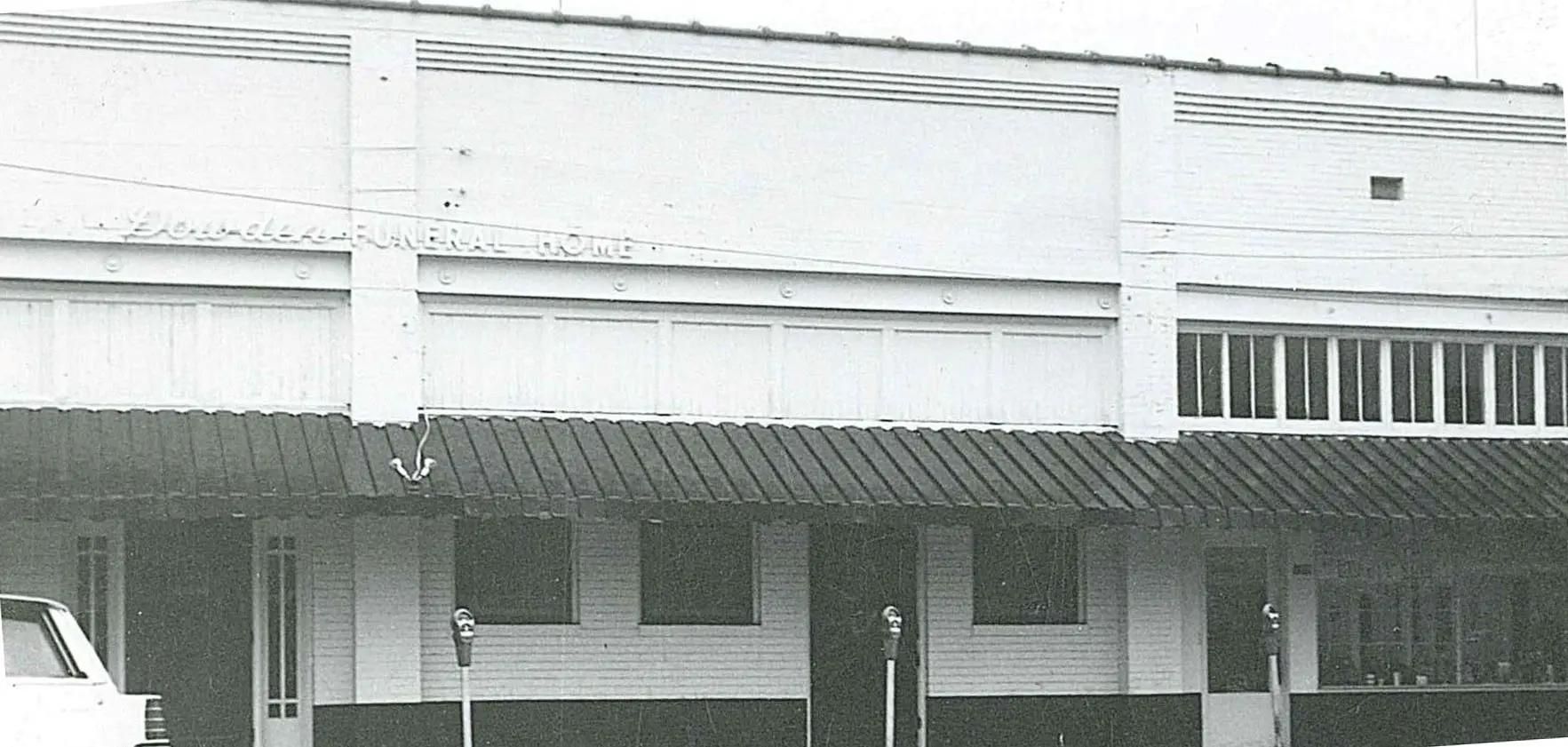 Black-and-white photo of a storefront with awnings, windows, and a parked vehicle at the left edge
