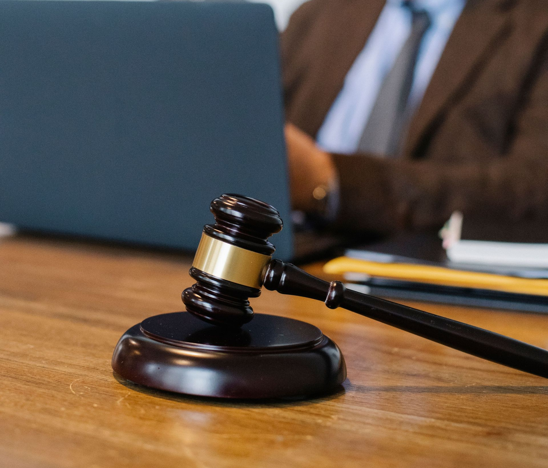 Judge’s gavel on a desk in a courtroom, with a blurred laptop and person in the background.