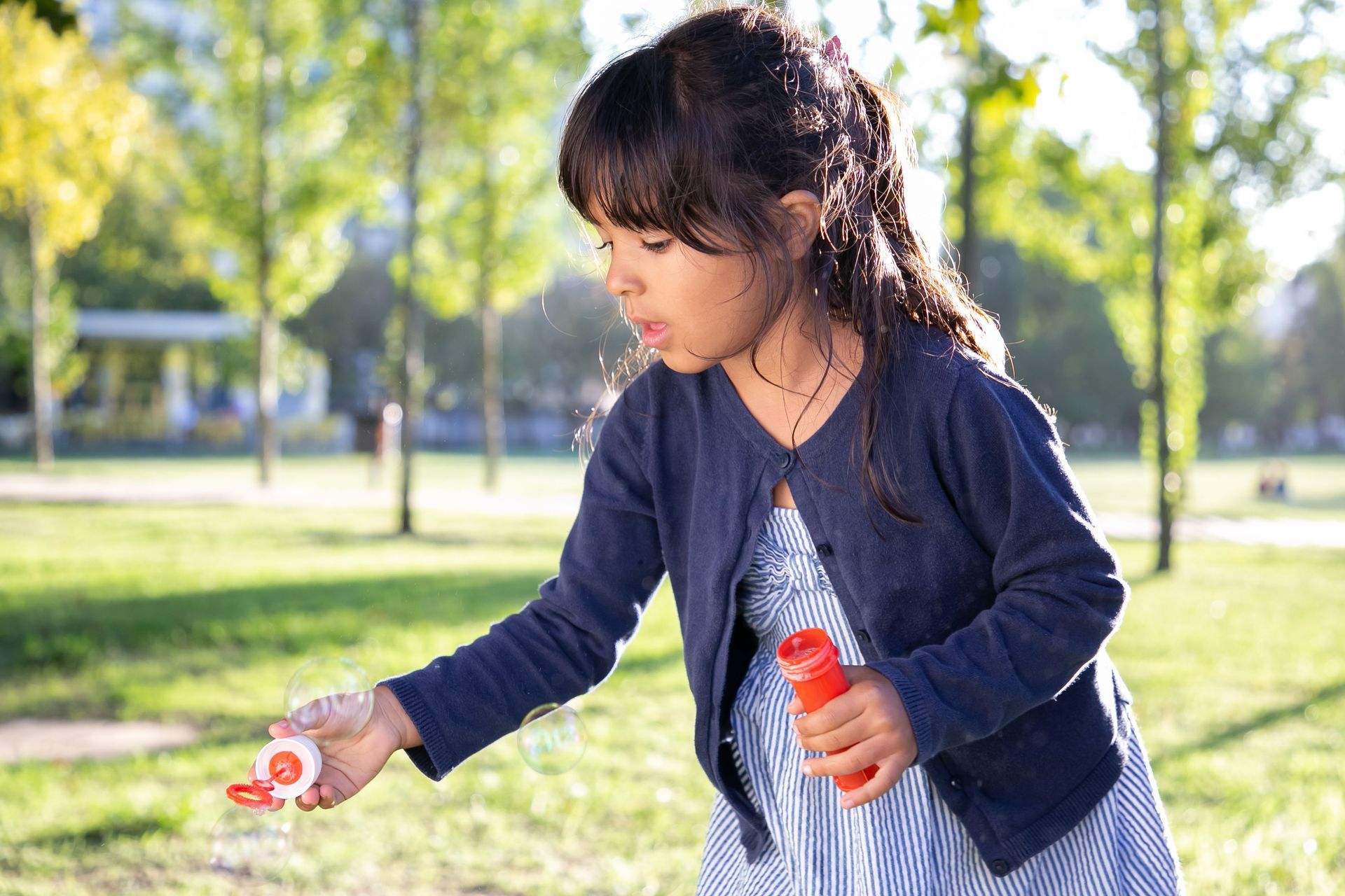 Woman in a blue jacket crouches in a park, holding a red bottle and reaching toward a small item on the ground.