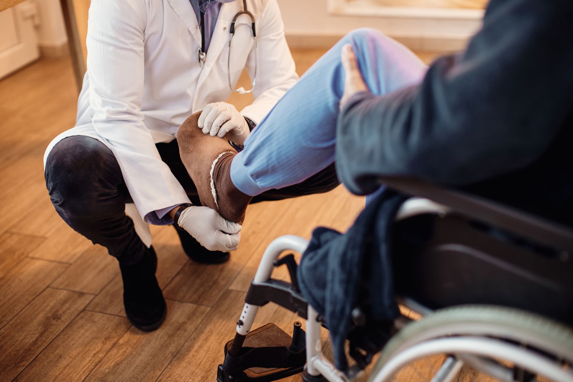 Physical therapist kneeling to examine a patient’s foot beside a wheelchair on a wooden floor
