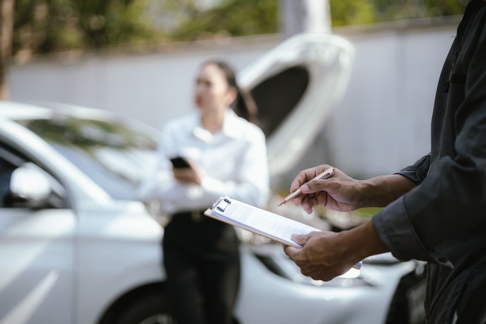 Person writing on a clipboard beside a white car, with another person standing nearby.