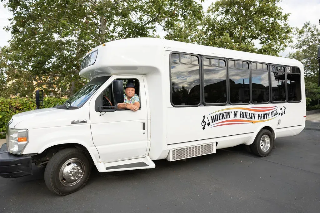 White minibus on a road with a driver visible, trees in the background