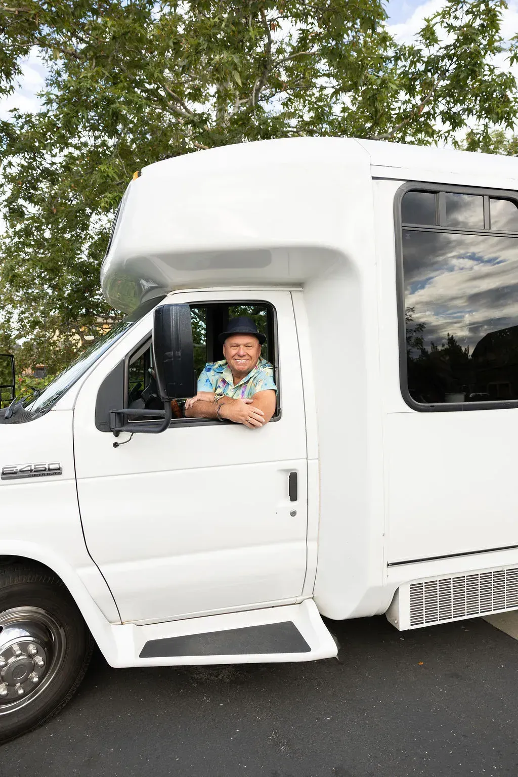 Man smiling from the driver’s seat of a white van parked outdoors under trees