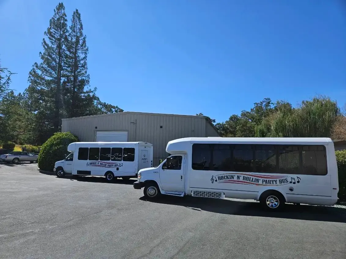 Two white shuttle vans parked in a sunny lot beside a large building and tall trees.