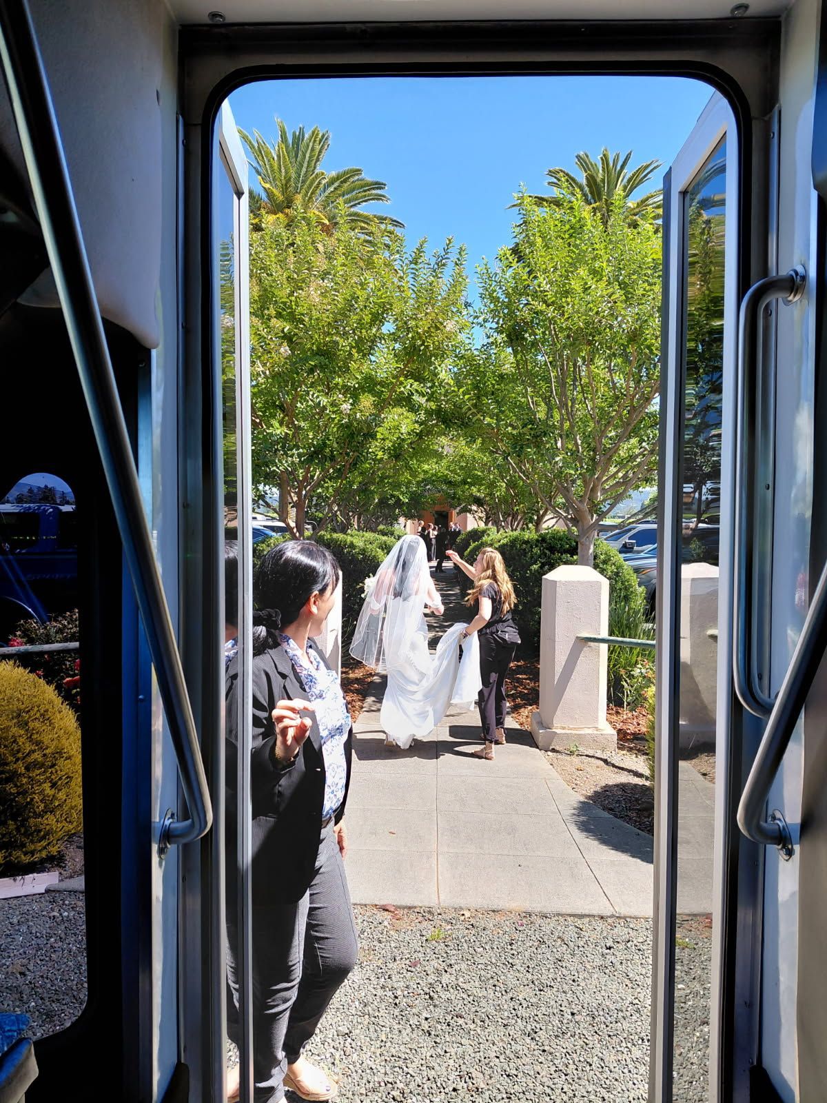 People in white robes walking along a sunlit path outside, seen through a vehicle doorway.