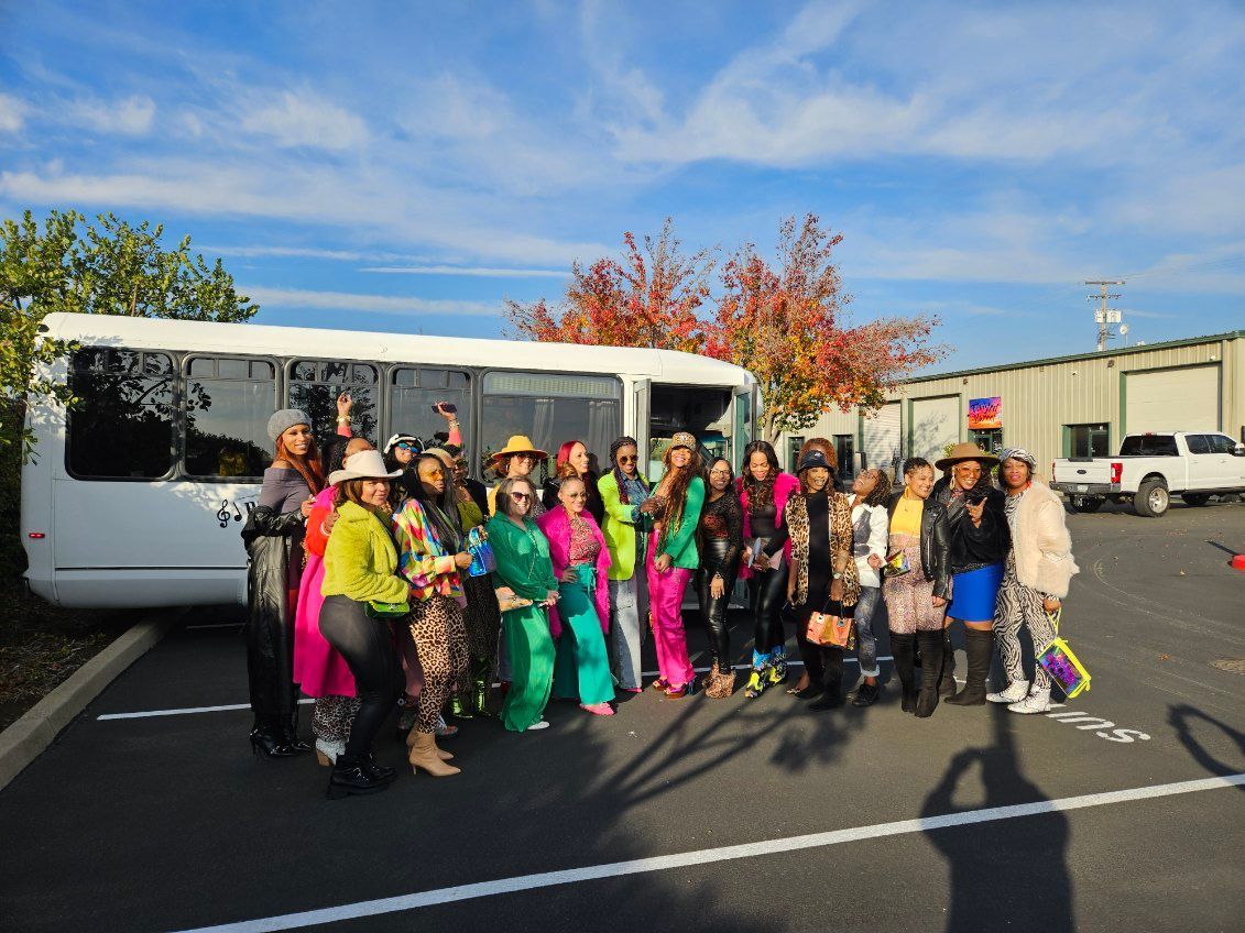 Group in colorful ski gear posing beside a white shuttle bus in a parking lot under a blue sky