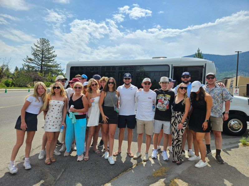 Group posing in front of a bus in a parking lot under a bright sky, with mountains in the background.
