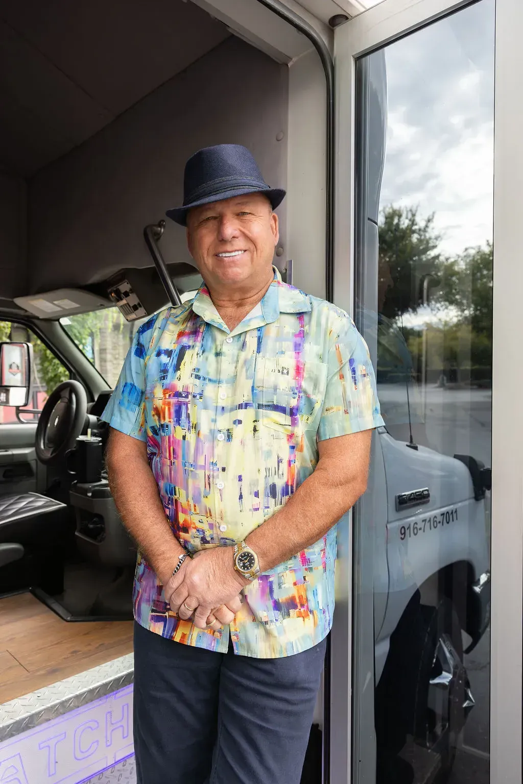 Man standing by a vehicle entrance, wearing a colorful shirt and dark hat, smiling at the camera.
