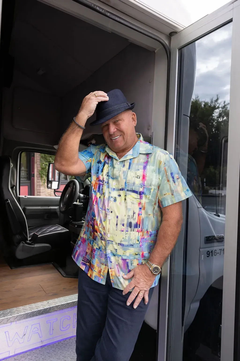 Man in a colorful shirt and hat standing in a van doorway, tipping his hat and smiling.