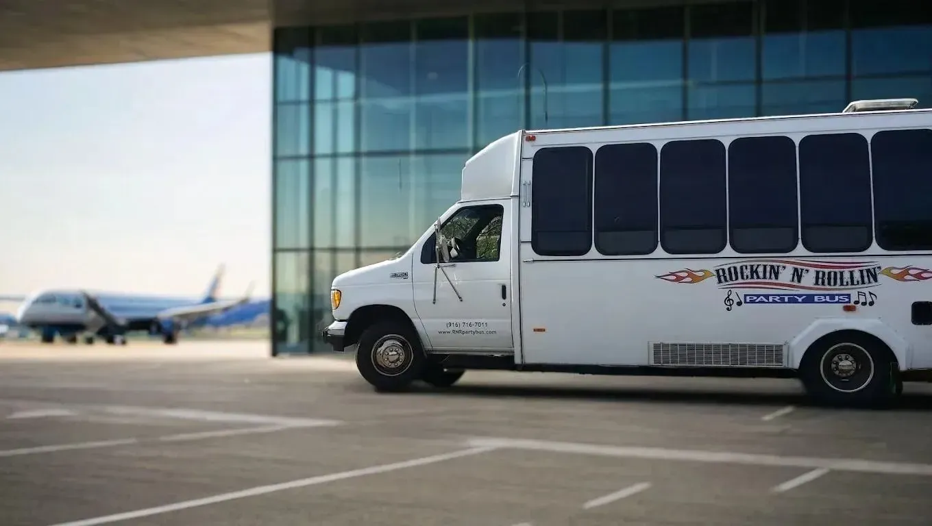 White shuttle bus parked outside a glass airport terminal, with an airplane in the background