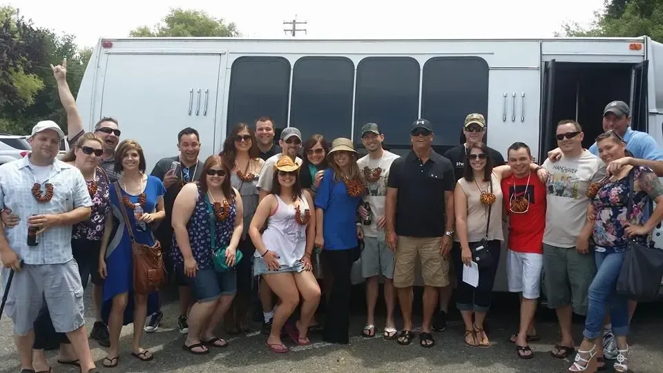Large group posing in front of a white bus on a street, wearing casual summer clothes and hats.