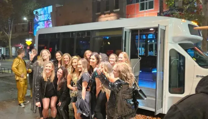 Group posing beside a white party bus at night, with one person in a gold outfit in the background.