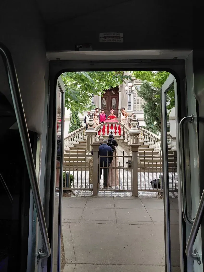 View through a vehicle doorway to a gated walkway, stone steps, and a red-robed statue in a leafy courtyard.