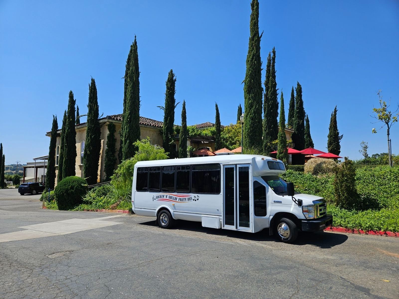White shuttle bus parked beside tall cypress trees and a hedge under a bright blue sky.