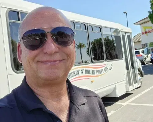 Smiling bald man in sunglasses taking a selfie beside a white shuttle bus in a sunny parking lot