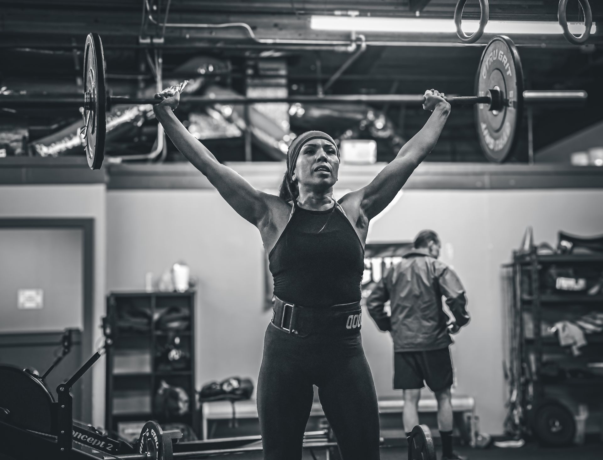 Woman lifting a barbell overhead in a gym, with a man in the background.
