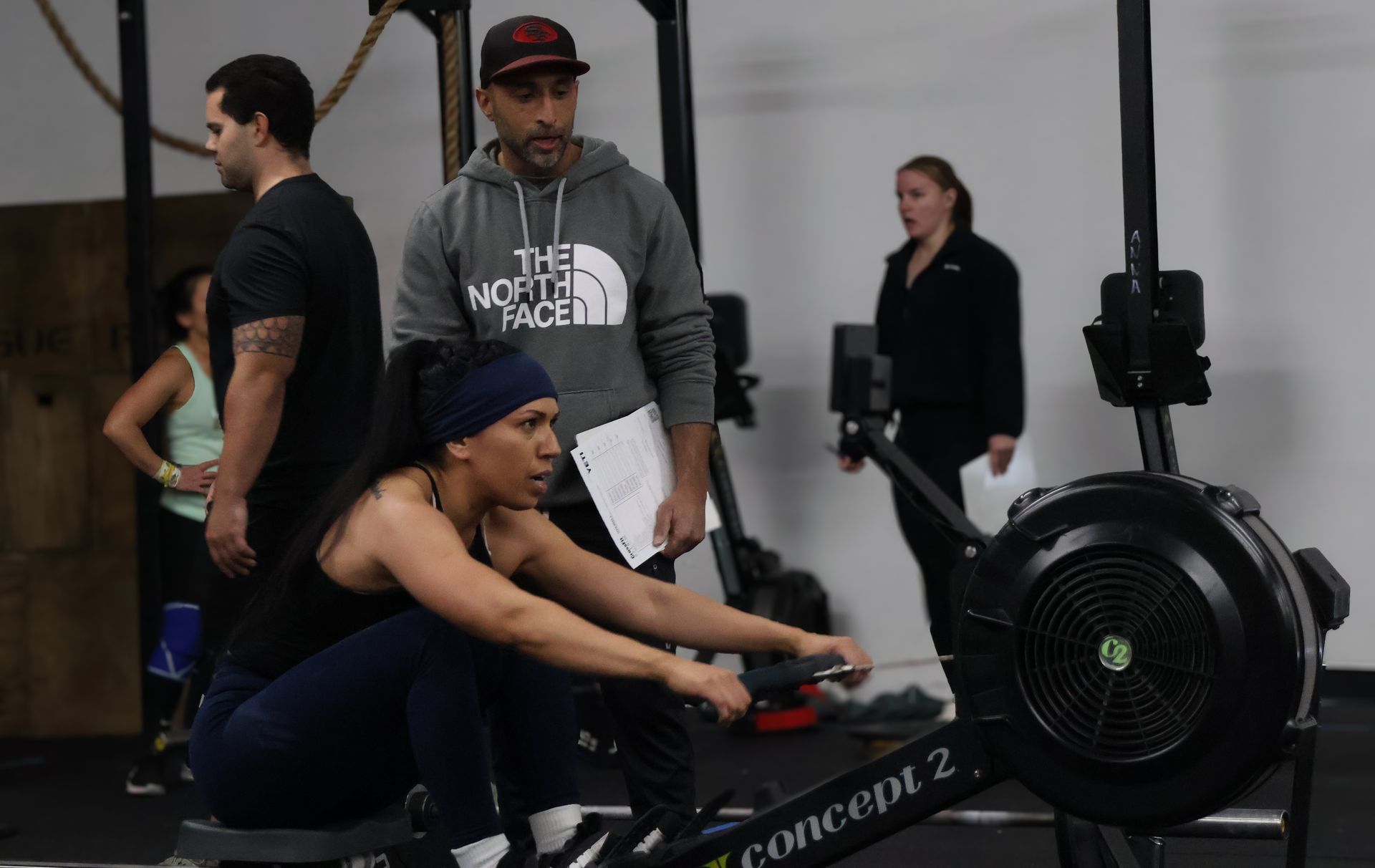 Woman rowing on a Concept2 machine in a gym while a coach observes nearby
