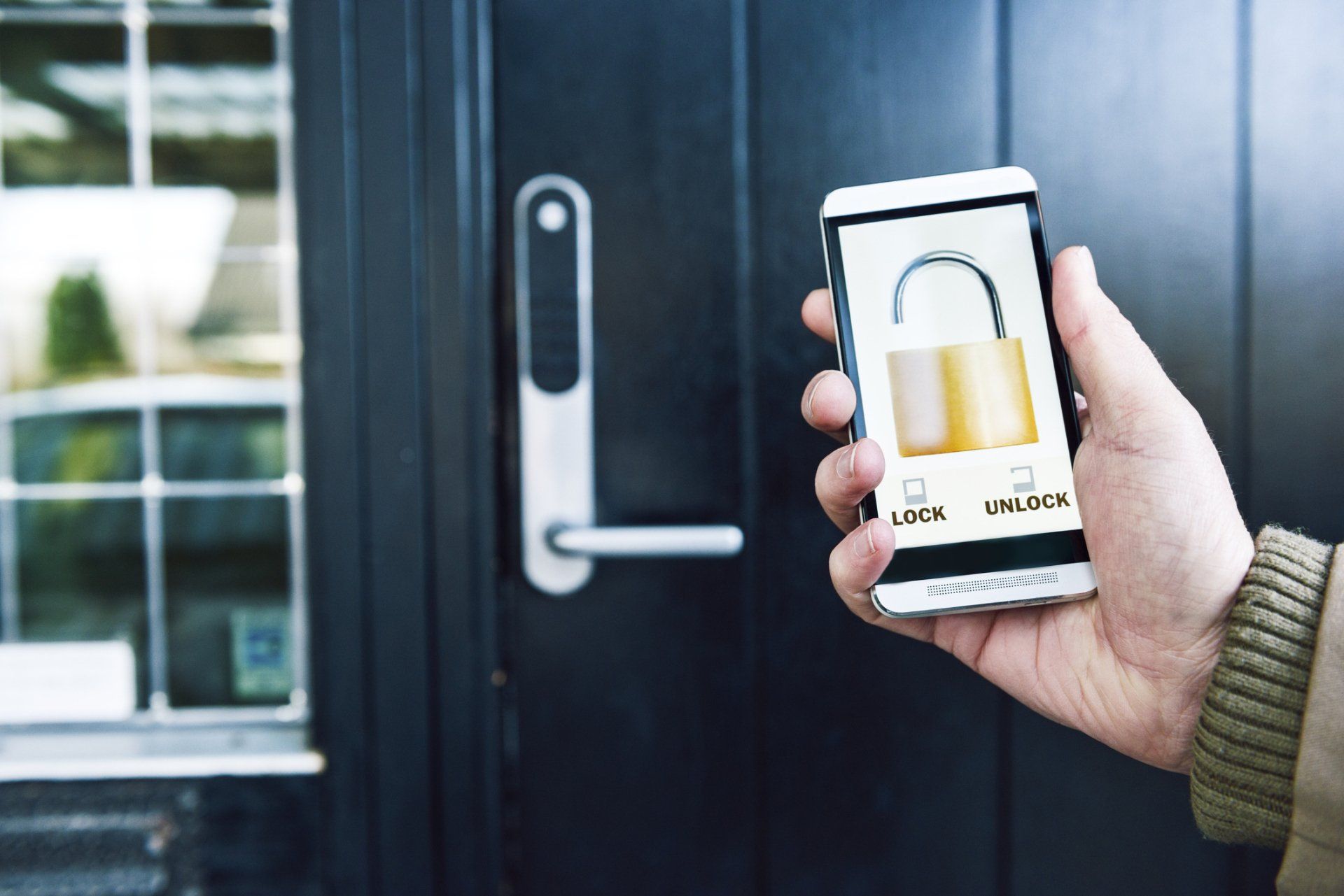 Person unlocks a door with a smartphone displaying a lock icon. The door is black with a silver handle.