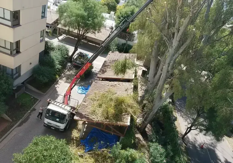 Una grúa retira escombros de un patio junto a un edificio de apartamentos, con árboles y lonas azules en las cercanías.