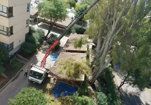 Una grúa retira escombros de un patio junto a un edificio de apartamentos, con árboles y lonas azules en las cercanías.