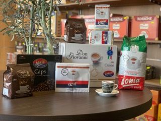 Vari prodotti da caffè Ionia su un tavolo rotondo, tra cui sacchetti, scatole e capsule, con una piccola tazzina da caffè espresso davanti.
