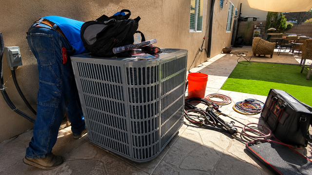 Worker bending over a large outdoor HVAC unit beside tools and cables in a narrow patio area
