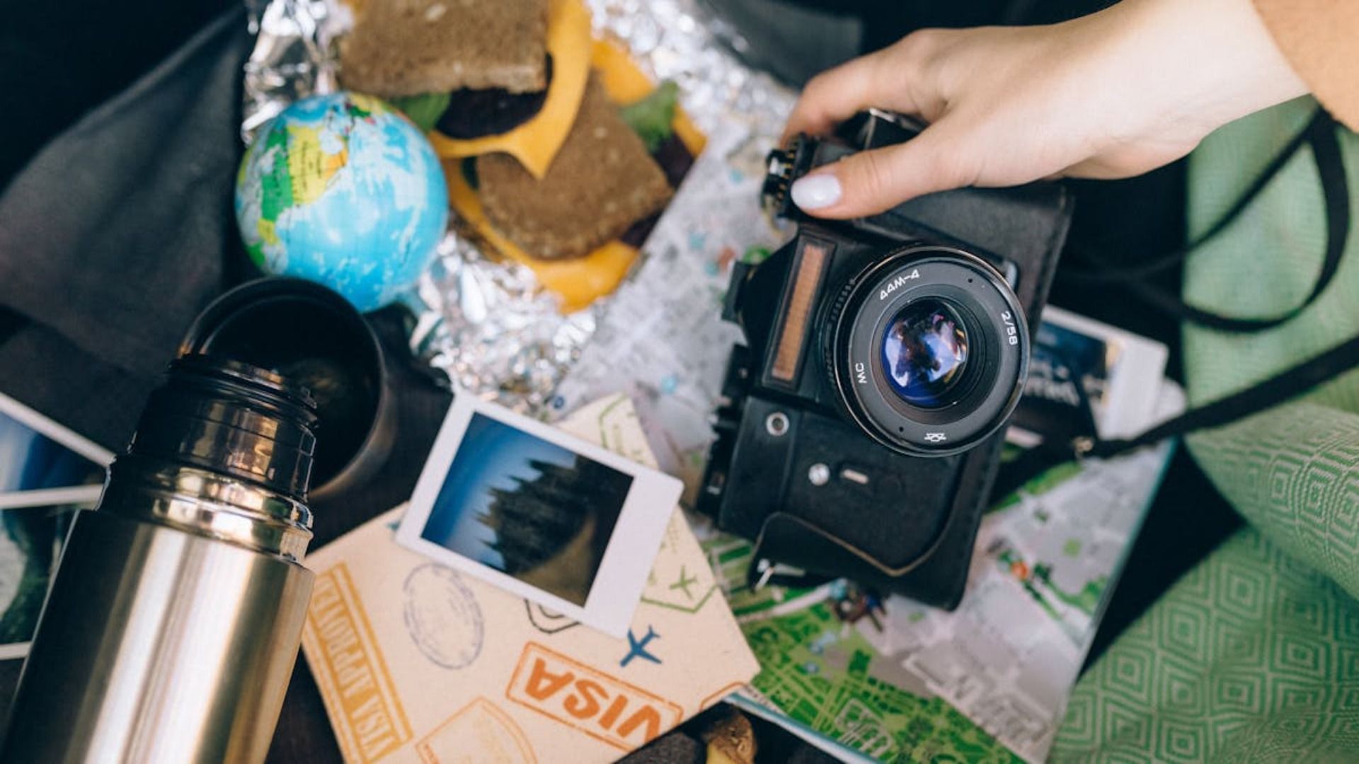 Mano sosteniendo una cámara, rodeada de artículos esenciales para el viaje: globo terráqueo, sándwich, fotos, termo y mapa.