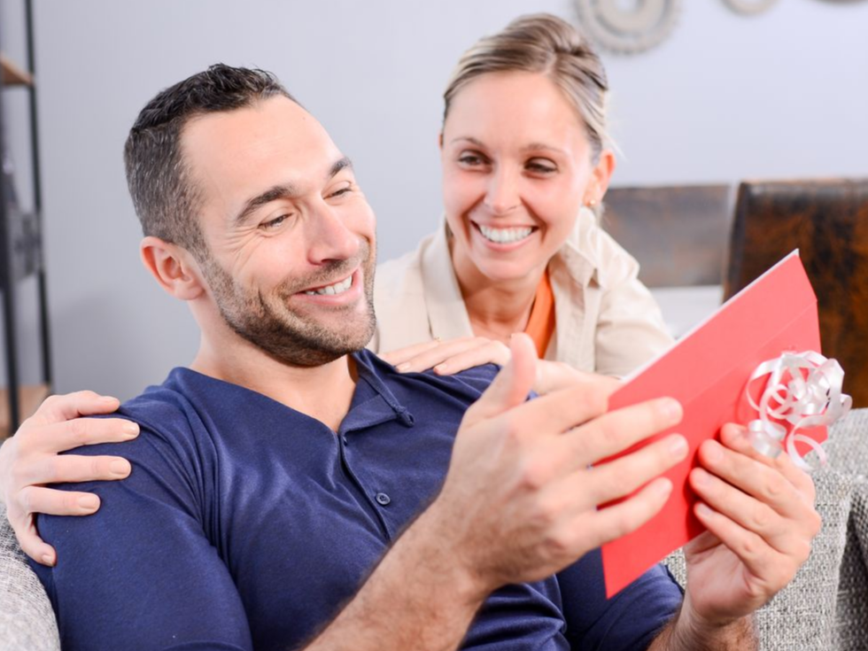 Lächelndes Paar auf einem Sofa, das eine rote Karte liest und ein kleines Geschenk in den Händen hält