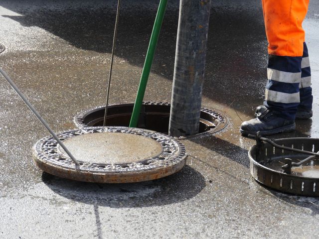 Person in orange workwear near open manhole and lid on wet pavement.