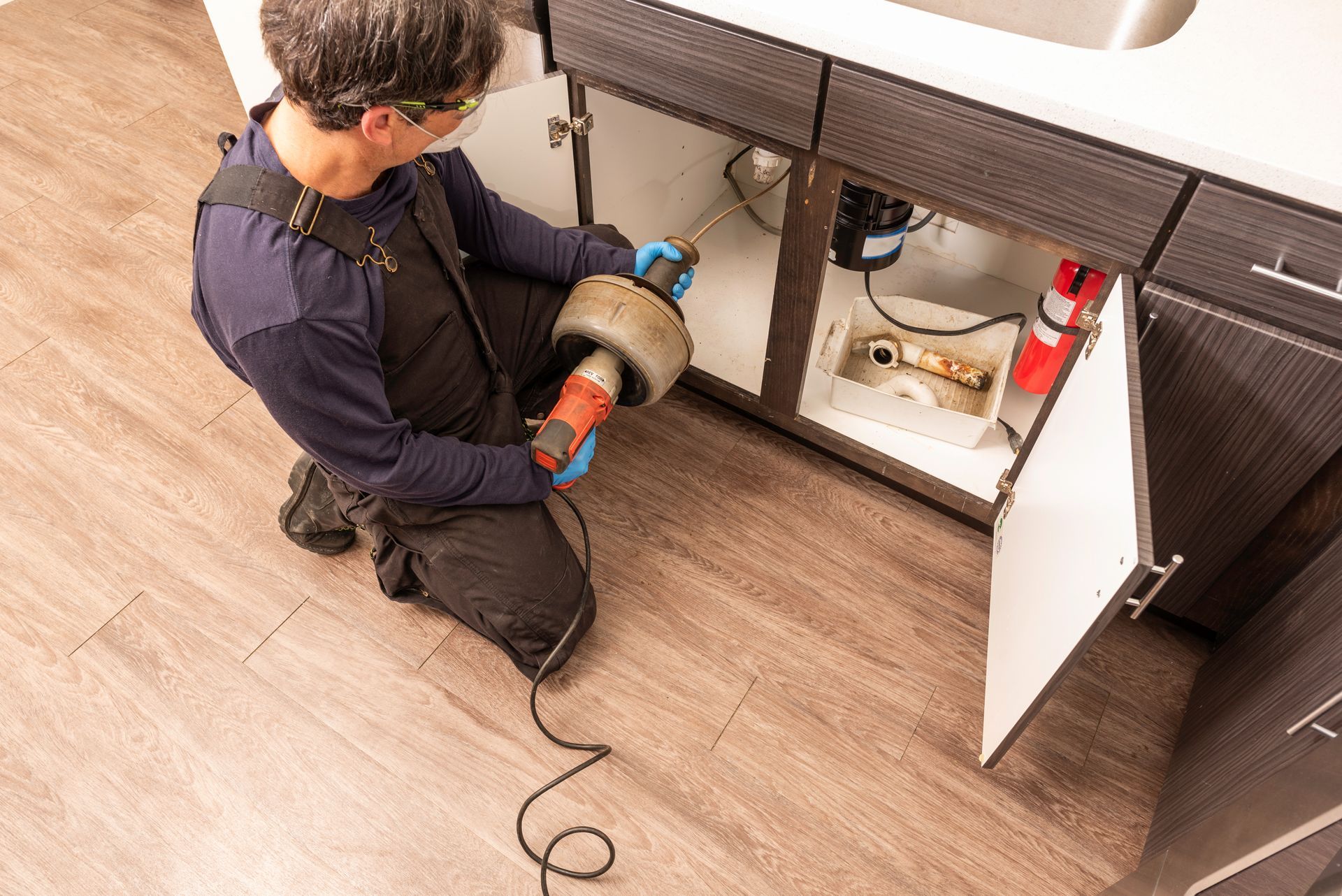 Plumber using a drain snake under a kitchen sink to unclog a drain. Plumber using a drain snake under a kitchen sink to unclog a drain.