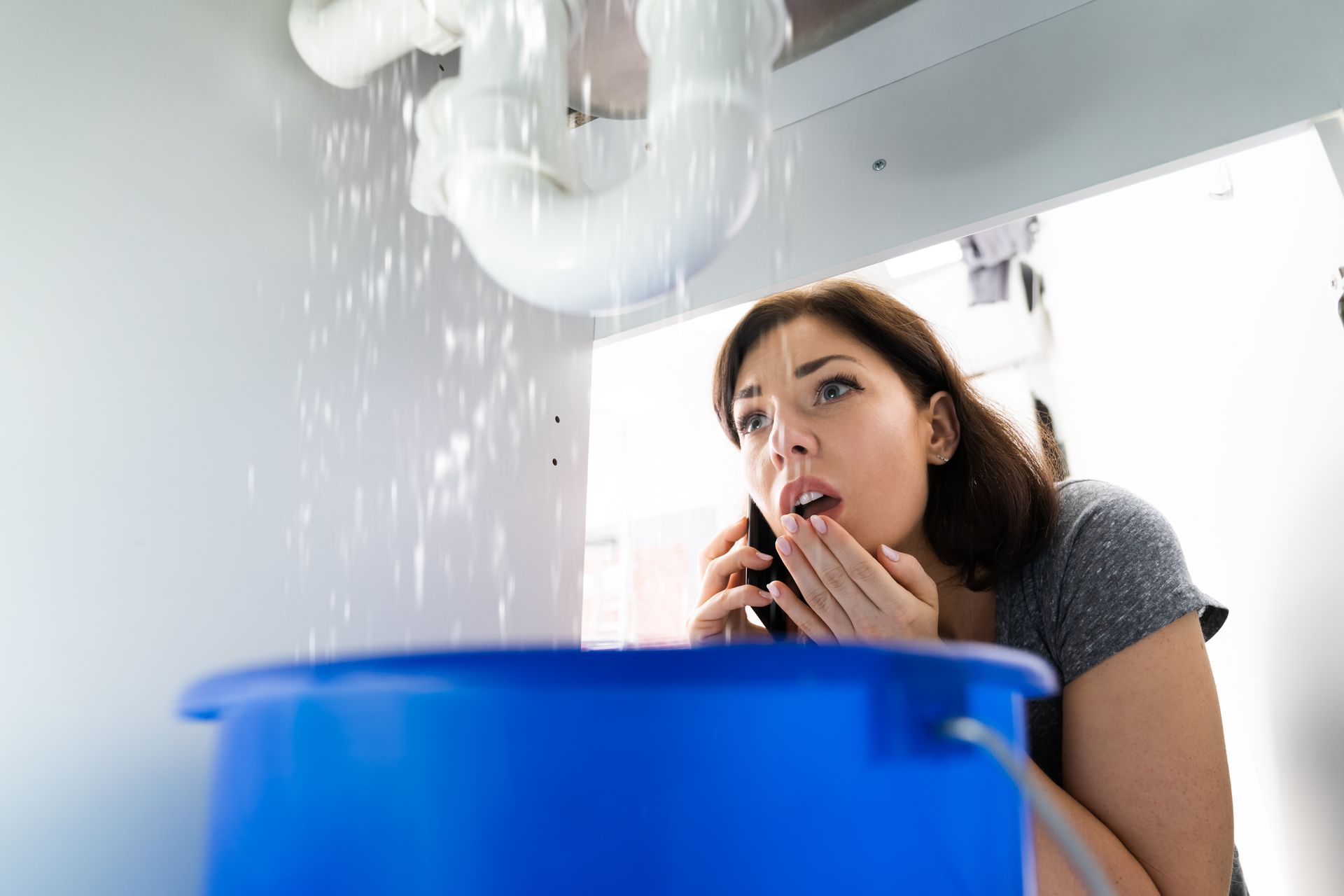 A worried woman calls a plumber as water leaks from the ceiling into a bucket.